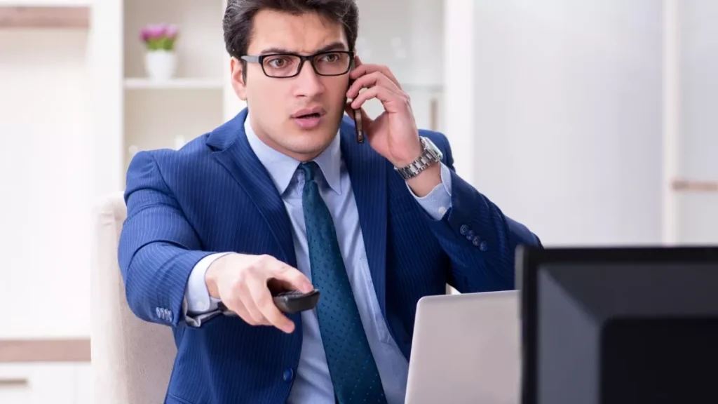 A business man using a phone and pointing a remote to a TV